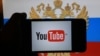 A hand holding a smartphone with YouTube's logo on the screen, in front of a computer screen in St. Petersburg, Russia showing the Russian national flag and coat of arms